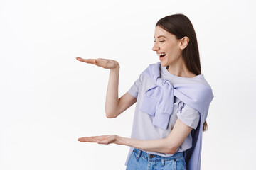 Cheerful girl looking at empty space, holding object box large size, laughing as staring at copyspace, standing over white background