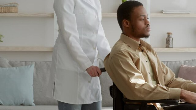 Nurse Pushing Wheelchair With Disabled Black Male Patient Indoors