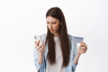 Online shopping. Brunette girl looks confused at smartphone screen, holding credit card, cant understand how register card to place an order, standing over white background