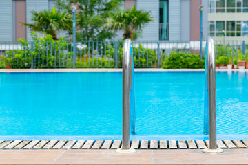 Swimming pool with stair and wooden deck in front with blurred green background. Outdoors on sunny day. Space for text. Poolside and outdoor spa vacation day.Grab bars ladder in the blue swimming pool