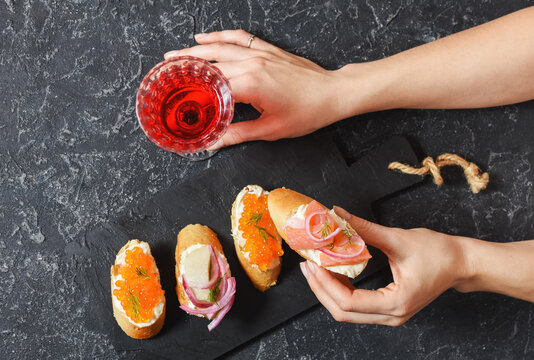 Female hands holds Bruschetta assortment with baguette, cream cheese, red caviar, salmon and glass of red wine on the stone background. Set of appetizers with various toppings