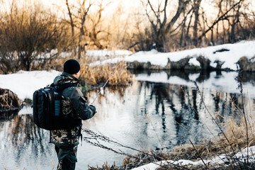 A fisherman with a fishing rod catches fish on the bank of a snow-covered river in early spring - Powered by Adobe