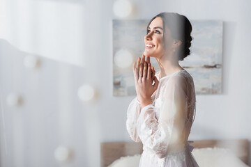 happy brunette bride looking away while holding hands near face on blurred foreground.