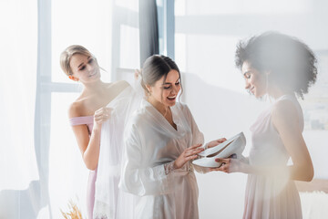 Naklejka premium amazed bride looking at white shoes in hands of african american bridesmaid, blurred foreground.