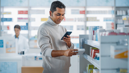 Naklejka na ściany i meble Pharmacy Drugstore: Portrait of a Handsome Young Indian Man Using Smartphone Device, Chooses to Purchase Best Medicine, Drugs, Vitamins. Shelves full of Sport Supplements, Health Care Products