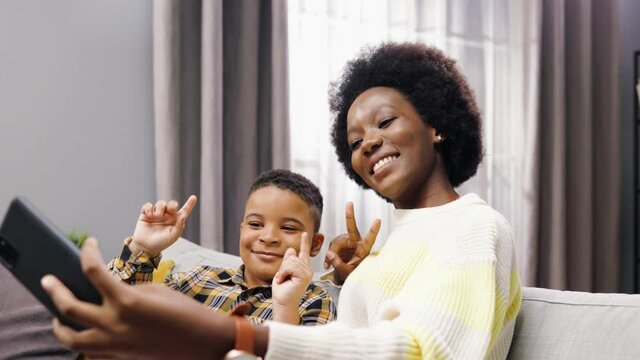 Close up of happy smiling African American young beautiful mom sitting on sofa with cute little son making gestures posing to cellphone camera at home taking selfie photos, leisure, family time