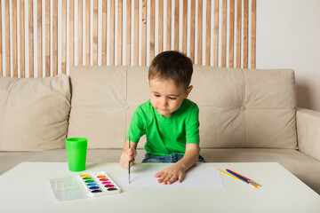Naklejka premium a little boy sits at a table and draws with a brush and paints on white paper in a room