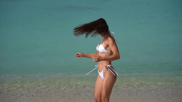 Beautiful fit woman in white bikini dancing in the beach on crystal aquamarine water. Concept of happy vacation in resort 