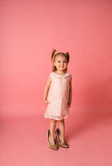 a cheerful little girl in a pink holiday dress stands in her mother's shoes on a pink background with a place for text
