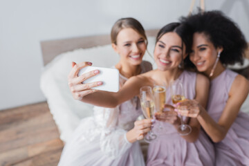 joyful woman taking selfie with bride and african american friend on blurred background.