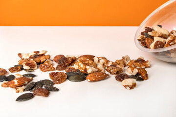 Fototapeta na wymiar Various nuts spilled from a cup on the white table against bright orange background. Selective focus, minimalistic, vivid colors.