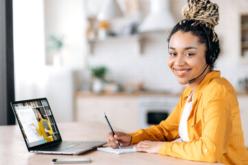 Distance learning by video call. Happy African American female student wearing headset, sits at work desk at home, studies remotely, looks at the camera, friendly smile,female teacher on laptop screen
