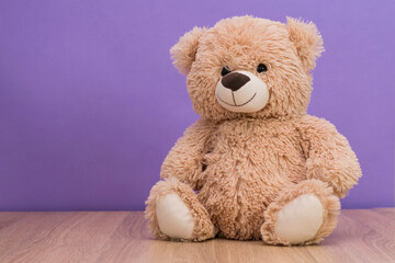Smiling teddy bear sits on the table against the background of a purple wall. Space for text, copy...