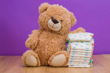 A teddy bear next to a stack of baby diapers. Baby toy teddy bear and diapers on the table against the background of a purple wall. Baby care, baby hygiene concept