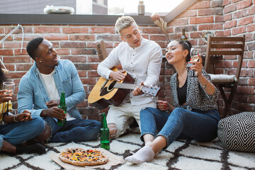 Multi ethnic group of people playing guitar and singing during rooftop party. Young hipsters...