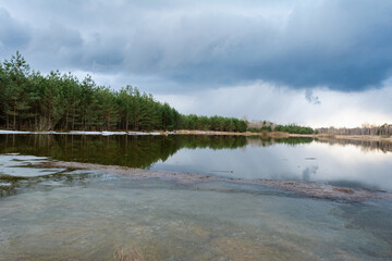 Forest lake with ice remnants, trees on the shore and dark rainy clouds in the sky. Wildlife spring...