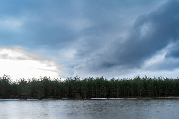 Forest lake with ice remnants, trees on the shore and dark rainy clouds in the sky. Wildlife spring landscape on cloudy evening