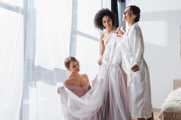 happy woman holding wedding dress near interracial bridesmaids.