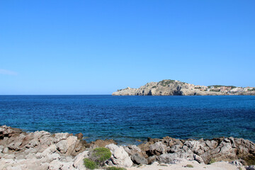 Rocky seashore on the island of Mallorca, in the distance you can see a gray stone island with buildings, a bush grows on the shore, clear sky, summer, sunny