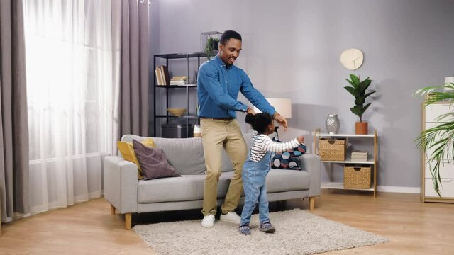 Joyful handsome young African American father with cute kid girl having fun at home in living room dancing, jumping and smiling in good mood. Little daughter playing with dad, leisure time concept