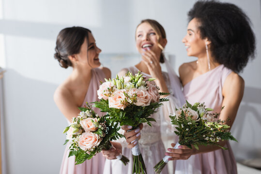 laughing bride and multicultural bridesmaids holding wedding bouquets on blurred background.
