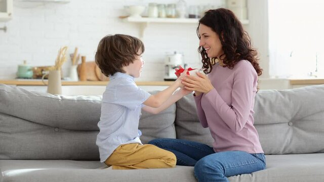 Happy mother sitting on sofa at home congratulating her kid giving surprise gift. Mom kissing child, receives wrapped gift box with red bow from son for her birthday. Loving family, holiday concept.