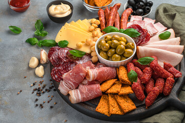 Round board with sausage and cheese slices, olives, basil and sauces on a dark gray background.