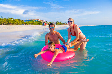 Family at the beach, in the ocean, mother and father holding pink lifesaver while daughter swims - obrazy, fototapety, plakaty