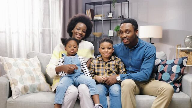 Camera approaching joyful African American family with kids sitting on sofa in room, looking at camera and smiling, positive emotions, parents with children gathered together at home parenting concept