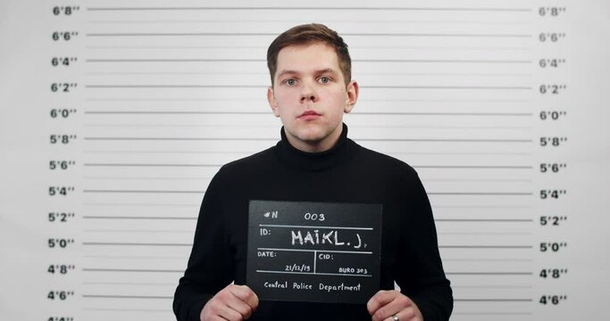 Portrait of guy in black turtleneck holding sign for photo in police department. Crop view of millennial man posing, raising head and looking to camera. Concept of crime.