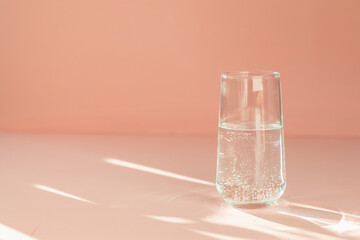 transparent glass on a peach background in the sunlight gives beautiful rays and glare in the reflection of the sun, a woman's hand takes the glass. stylish photo of fresh clear water in a glass