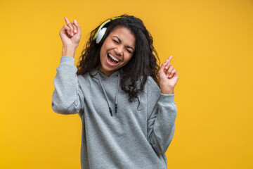 Portrait of dark skinned hipster girl wearing grey basic hoodie and wireles headphones enjoying listnening to music and dancing isolated over bright colored orange yellow background