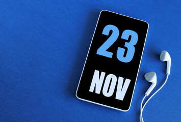 November 23. 23 st day of the month, calendar date. Smartphone and white headphones on a blue background. Place for your text. Autumn month, day of the year concept