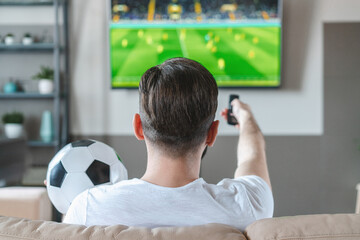 Casually dressed man spending his day-off at home sitting on sofa watching soccer play live braodcast on tv and supporting his favourite team