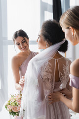 Naklejka premium smiling bridesmaids preparing african american bride for wedding, blurred foreground.