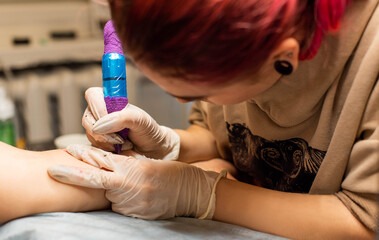 Charming girl with pink hair and tattoos stuffs a tattoo on a woman's hand in a tattoo parlor,...