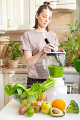 happy and cheerful caucasian woman in a t-shirt in her kitchen makes a green smoothie from fresh fruits and vegetables, a healthy vegan and vegetarian drink for health and proper nutrition after