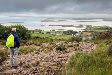 Fototapeta na wymiar Bald male tourist with backpack looking at beautiful scenery. Westport, county Mayo, Ireland. Foot path to Croagh Patrick. Irish nature and travel concept