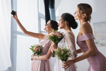 african american bride with bridesmaids holding wedding bouquets while taking selfie on smartphone. - obrazy, fototapety, plakaty