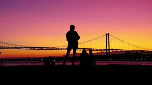 Girl take pictures of young couple sitting on pier against beautiful sunset afterglow, silhouetted shot. Scenic evening at bank of Tagus river, large suspension bridge seen on background