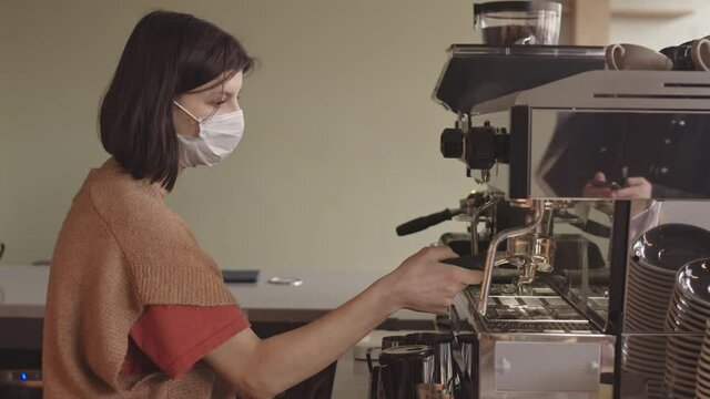 Medium shot of young female bartender wearing casual clothes and mask standing in bar near automatic coffee machine, cleaning it and preparing for making next cup of coffee