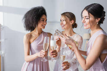 smiling woman pouring champagne near happy bride and african american bridesmaid.