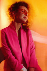 A beautiful, well-groomed girl in a crimson jacket and glasses sits on a chair against a background of colorful flying fabric.