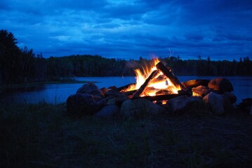 camp fire burning in a fire pit next to the lake - obrazy, fototapety, plakaty