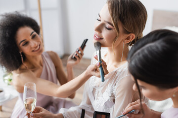 young bride holding champagne glass while multicultural bridesmaids applying makeup, blurred background.