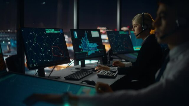 Female Air Traffic Controller with Headset Talk on a Call in Airport Tower at Night. Office Room is Full of Desktop Computer Displays with Navigation Screens, Airplane Flight Radar Data for the Team.