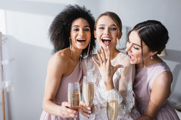 excited woman showing wedding ring near interracial bridesmaids with champagne glasses.