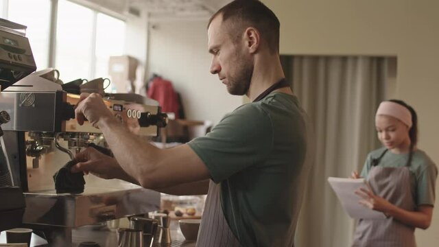 Lockdown of middle-aged Caucasian man wearing uniform standing at coffee machine in cafe, cleaning it and then coming to his female colleague writing in notepad on background