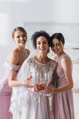 joyful african american bride holding champagne near friends in bedroom.