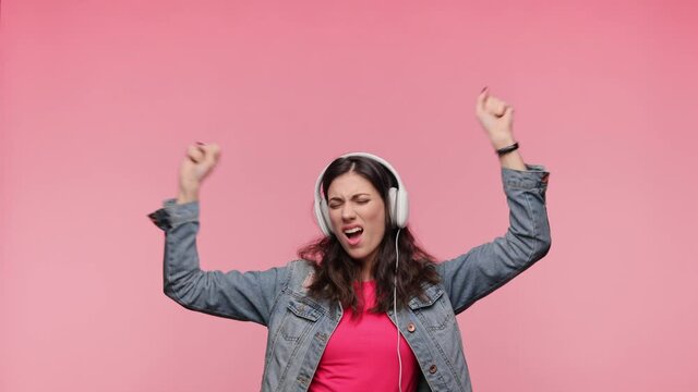Brunette Woman in casual denim jacket pink t-shirt isolated on pink background in studio. People lifestyle concept. Girl listen music in headphones dancing fooling around have fun gesticulating hands.
